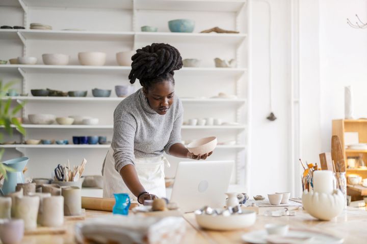 A woman wearing an apron and a grey turtleneck is doing pottery in a pottery studio.