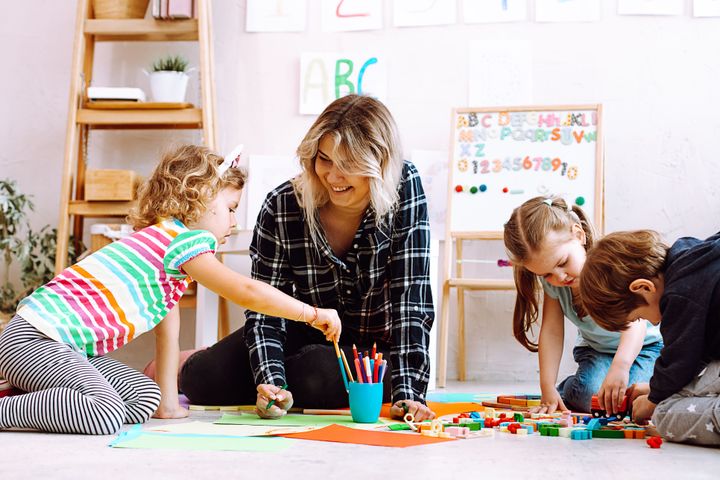 A young female teach is working with 3 children in a classroom. They are colouring on the floor and have art behind them.